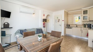 A compact living space with a dining table, wicker chairs, a sofa, a TV, and a kitchen area.