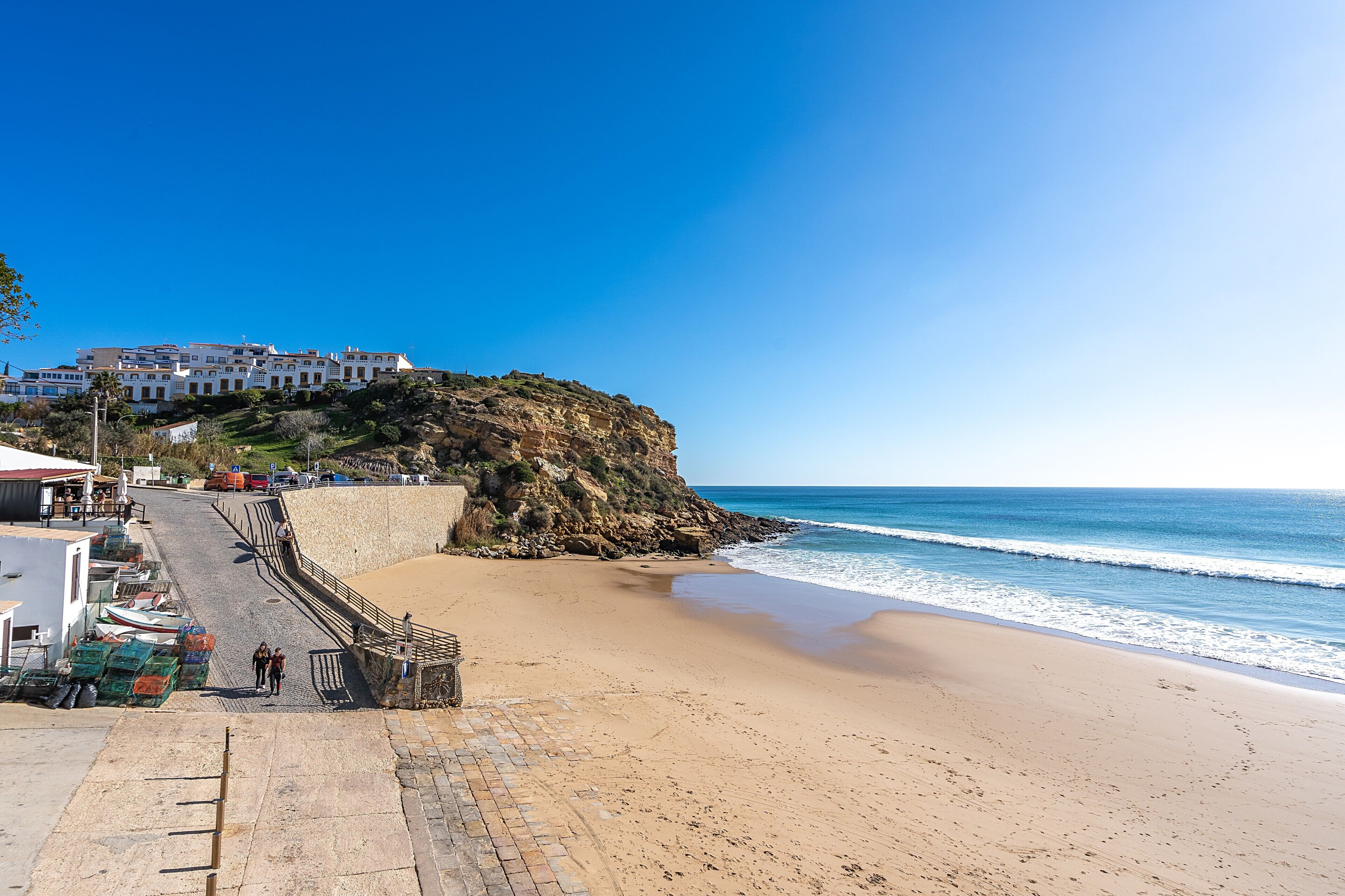 Un pueblo costero con una playa, una escalera que lleva a un edificio en la cima de un acantilado, y un cielo azul despejado.