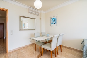 Uma sala de jantar com uma mesa de vidro, quatro cadeiras, uma luminária no teto, um ar condicionado e um quadro na parede.