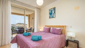 A bedroom with a bed, a bedside table, a lamp, a window with curtains, and a view of the outside. - Apartment 'Brightness Comfort In Burgau' with Sea View, Wi-Fi and Air Conditioning (Burgau)