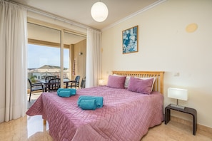 Quarto com cama, mesa de cabeceira, luminária, janela com cortinas e vista para o exterior.
