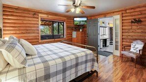 Habitación tipo cabaña de madera con cama, televisor, ventilador de techo y vistas del exterior a través de una ventana.