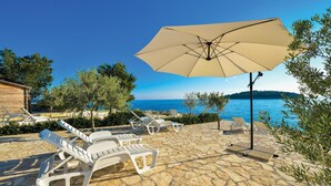 A stone patio with white lounge chairs and a beige umbrella, overlooking a body of water and a distant island. - Stunning apartment in Rogoznica (Rogoznica)
