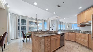 A modern kitchen with a central island, granite countertops, wooden cabinets, and a large window offering a view of the balcony. - Mediterranean West 601W | Next to Florabama | Heated Indoor Pool w/Swim-Thru | My Beach Getaways (Pensacola)