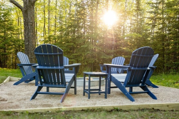 A forested area with four Adirondack chairs and a small table.