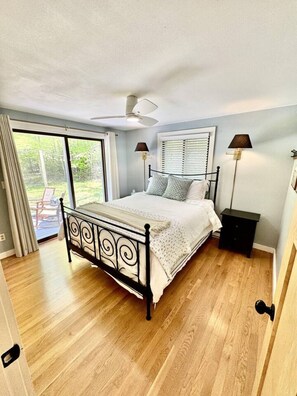 A bedroom with a bed, a ceiling fan, a window with blinds, a nightstand, and a view of the outdoors.