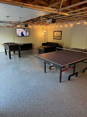 A room with a foosball table, ping pong table, and a TV screen.