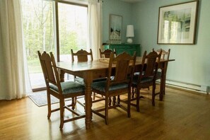 A dining room with a wooden table and chairs, a window with curtains, a lamp, and a painting on the wall.