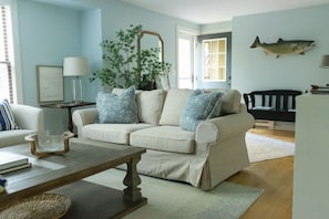 A living room with a beige sofa, a coffee table, a lamp, a mirror, a plant, and a fish wall decoration.