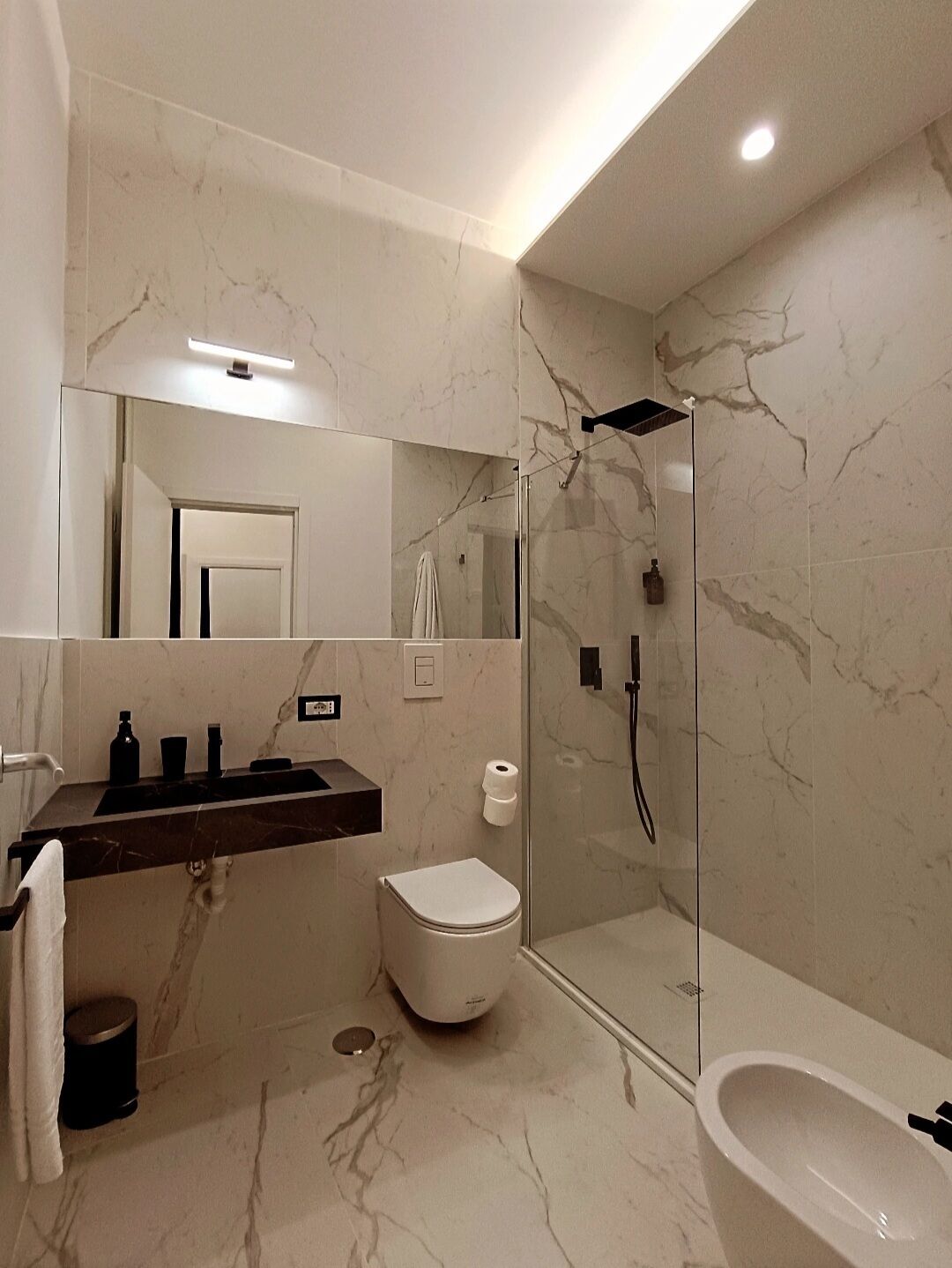 A modern bathroom with a marble floor, a glass-enclosed shower, a white toilet, and a black sink with a mirror above it.