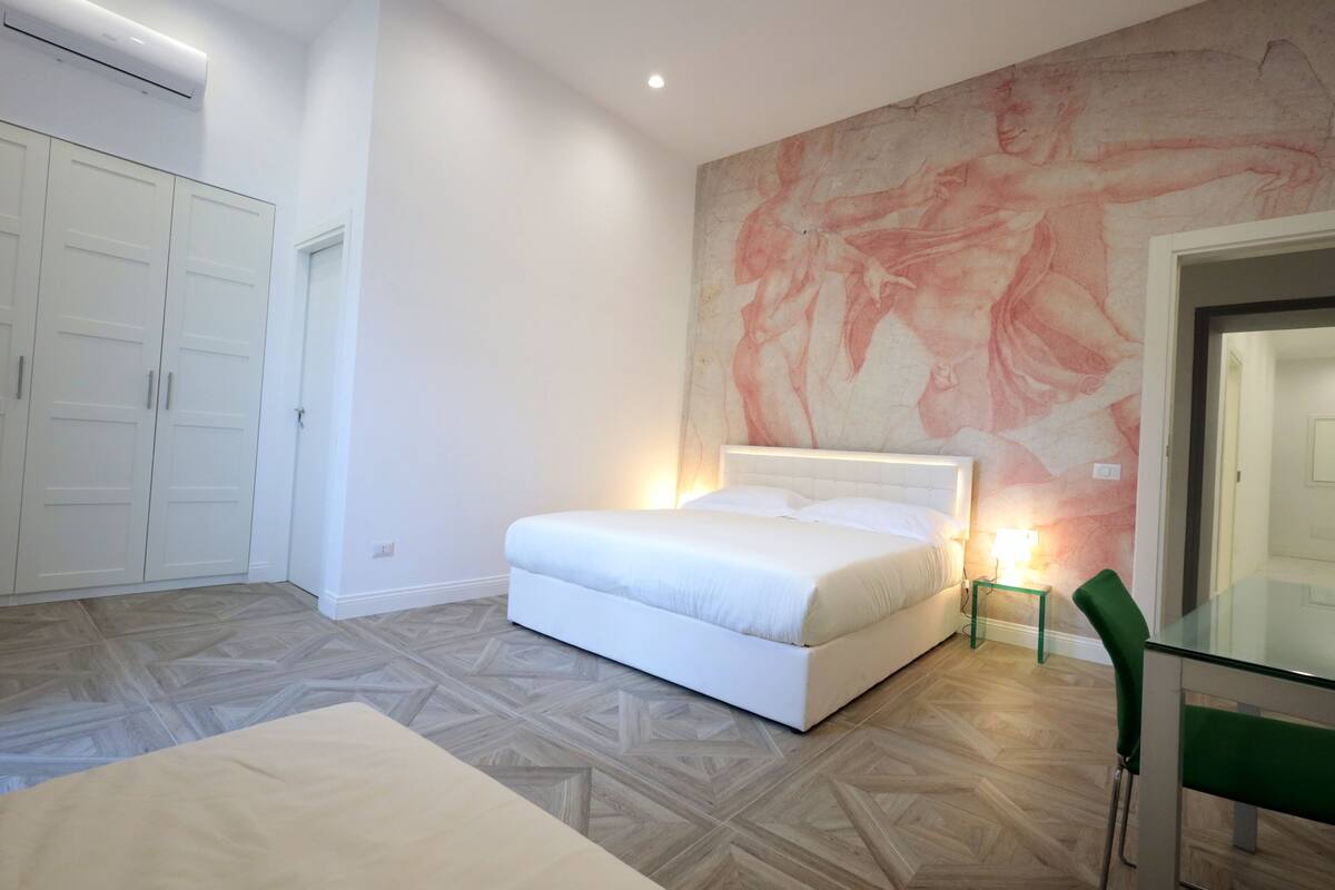 Ein Schlafzimmer mit einem Bett, einem grünen Sessel, einem Glastisch und einem großen Wandgemälde.