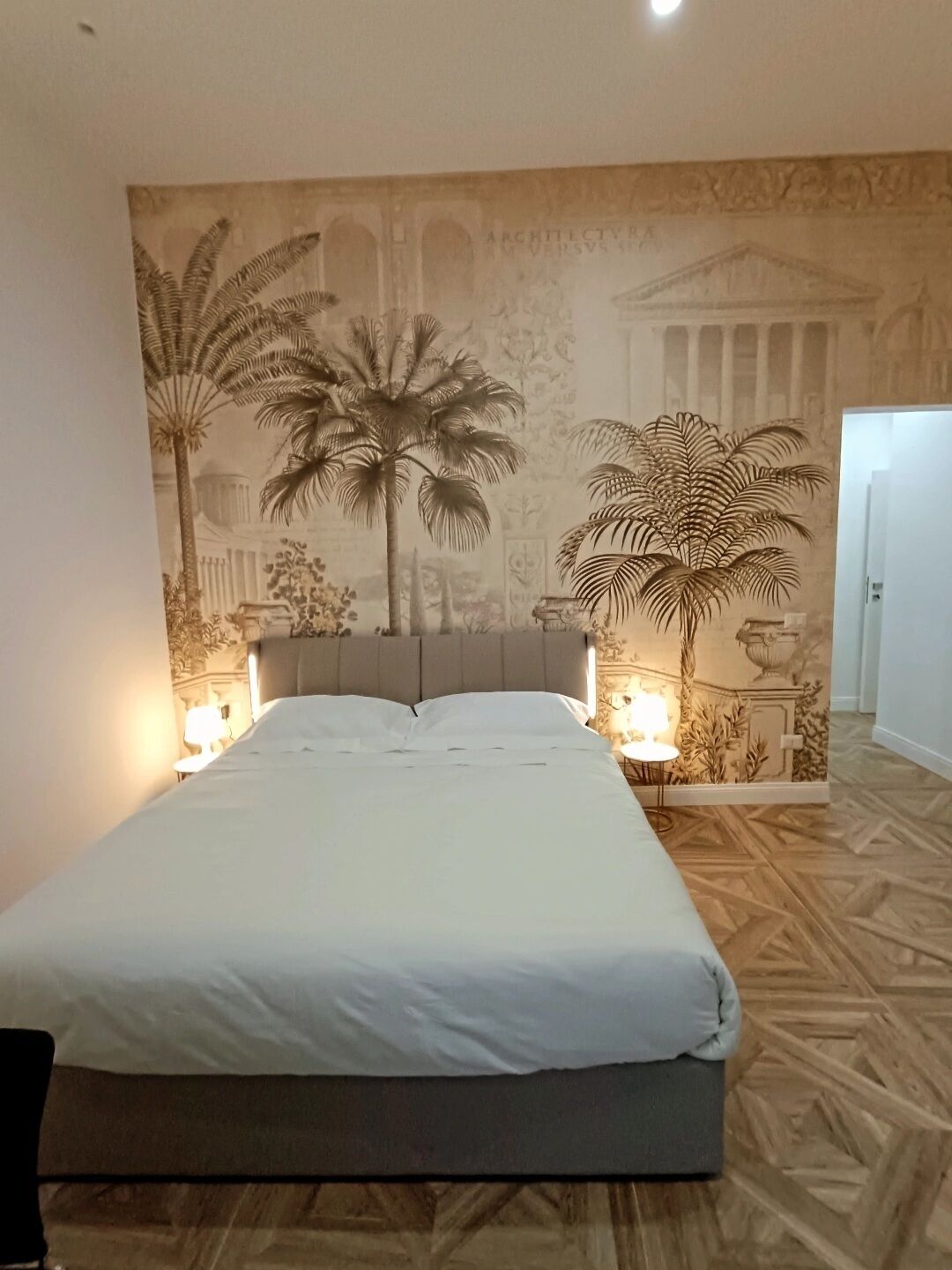 Camera da letto con un letto grande, due lampade sui comodini e un murale raffigurante palme e architettura classica.