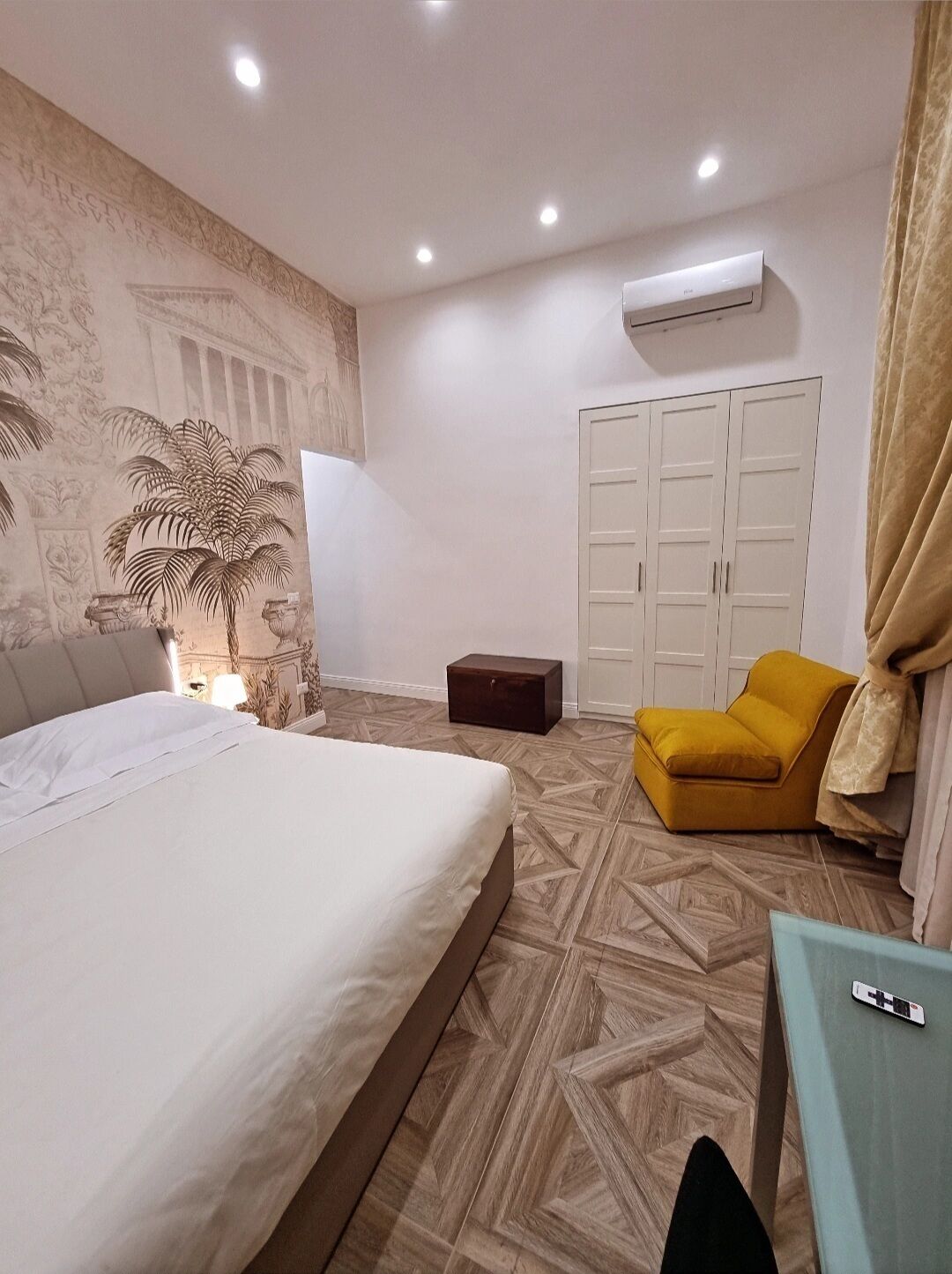 A hotel room with a bed, a small table, a yellow armchair, a wardrobe, and an air conditioning unit.