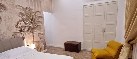 A hotel room with a bed, a small table, a yellow armchair, a wardrobe, and an air conditioning unit.