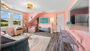 A modern living room with a pink wall, a wooden floor, a sofa, a chair, a lamp, a TV, and a kitchen area with a fridge.