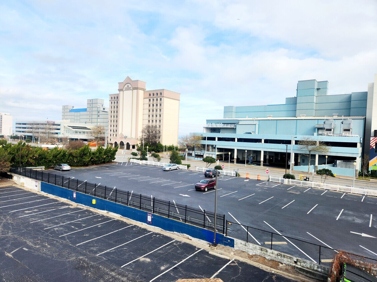 A parking lot with multiple rows of parking spaces, a blue fence, and a building with a blue roof in the background.