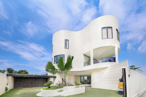 A modern, white, multi-story house with a curved facade, large windows, and a well-maintained garden with palm trees. - Villa Alinea (Tabanan)