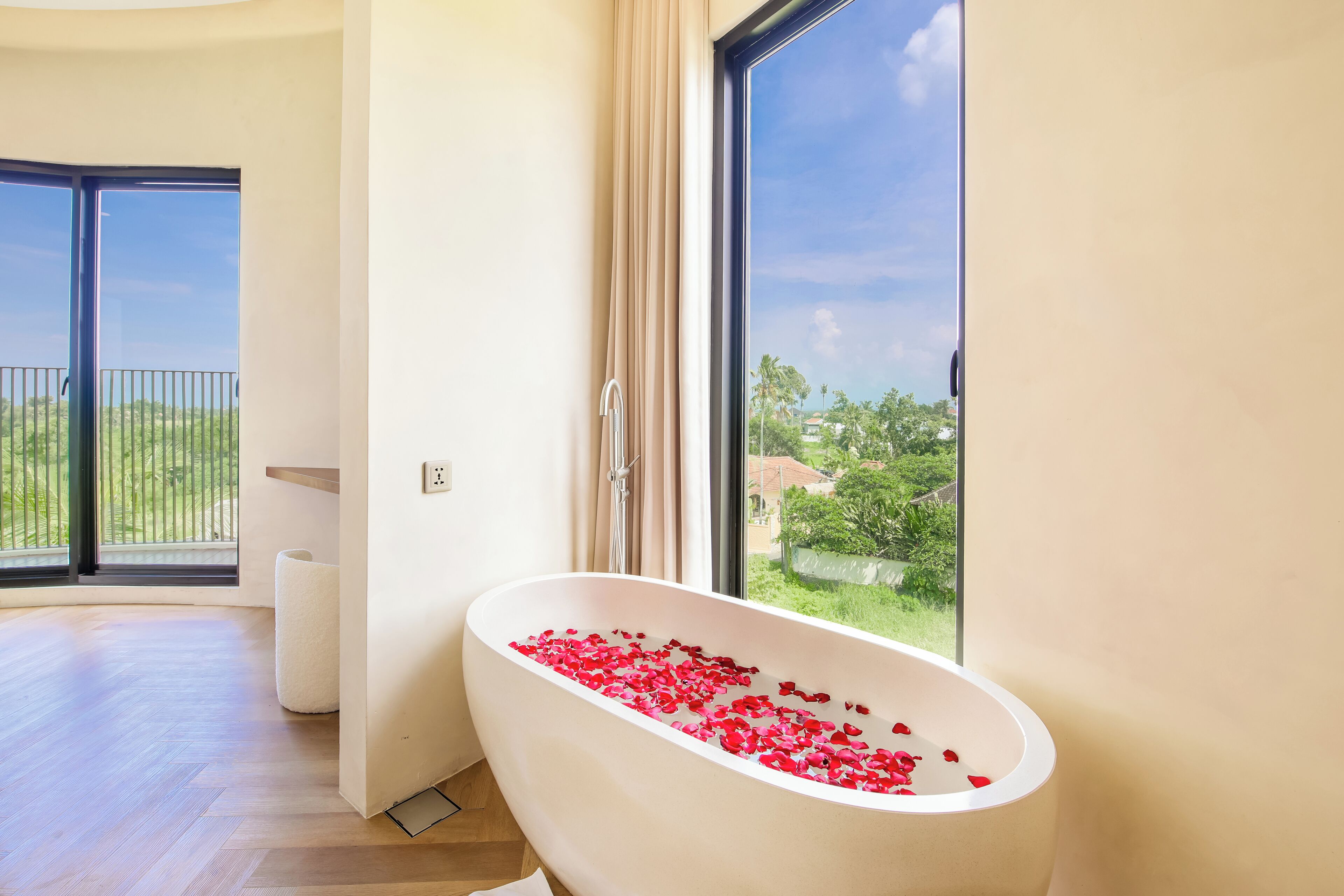 A modern bathroom with a freestanding bathtub filled with rose petals, large windows offering a view of greenery and a balcony.