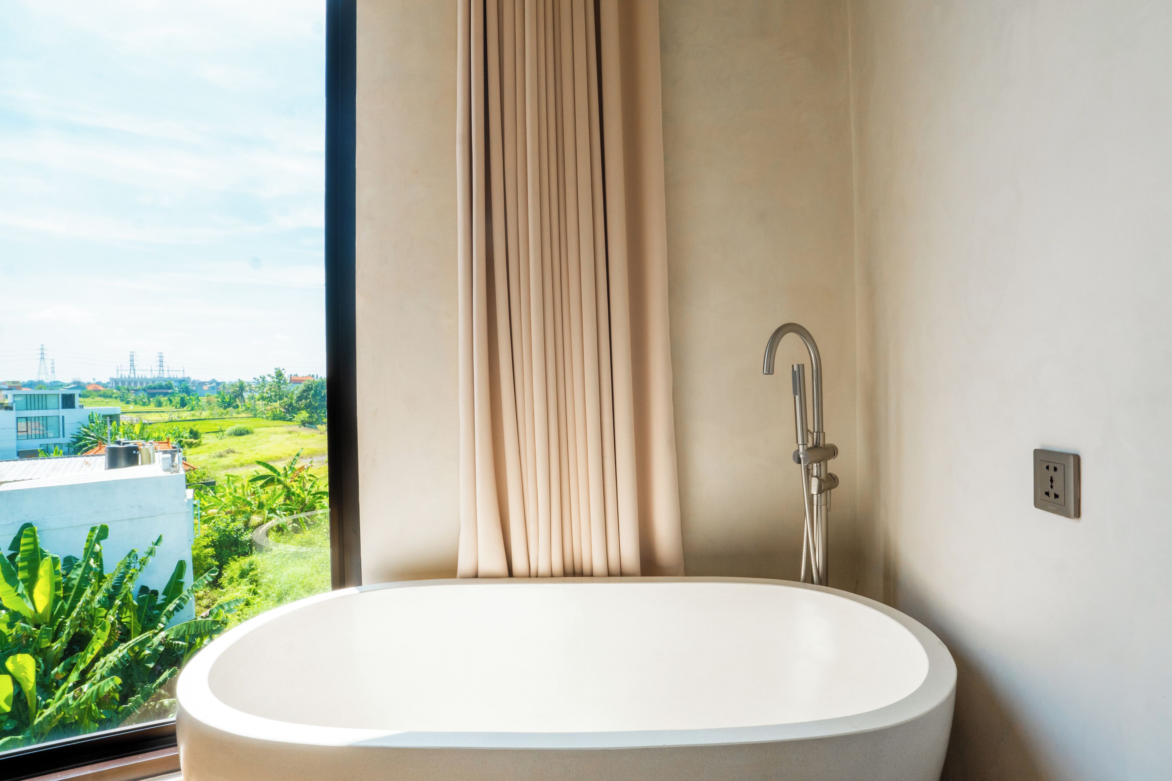 A modern bathroom with a large oval sink, a visible showerhead, and a view of greenery and buildings outside the window.