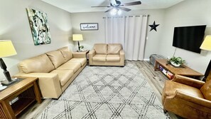 Uma sala de estar com sofá, poltrona e mesa de centro. Há um ventilador de teto, uma TV e um quadro na parede.