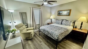 A bedroom with a bed, a chair, a nightstand, a lamp, a ceiling fan, and a window with curtains.