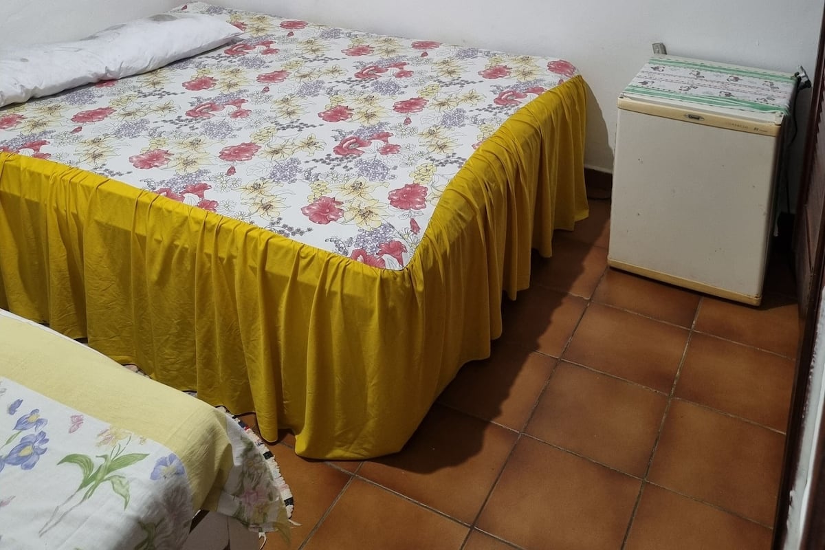 Ein kleines Zimmer mit einem Einzelbett, einer gelben Tagesdecke, einem kleinen Nachttisch und einem Kühlschrank. An der Wand hängen drei gerahmte Bilder.