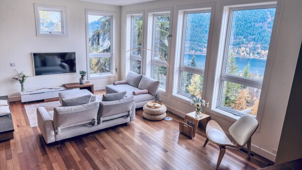 A modern living room with a sofa, a TV, and large windows offering a view of a forest and lake. - NEW Modern Mountain Haven | Luxe View, Sauna, Hot Tub & Fireplace (Nelson)