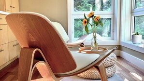 A modern interior with a wooden chair, a vase with flowers, and a view of a forest through a large window. - NEW Modern Mountain Haven | Luxe View, Sauna, Hot Tub & Fireplace (Nelson)
