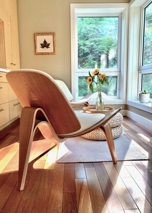 A modern interior with a wooden chair, a vase with flowers, and a view of a forest through a large window.