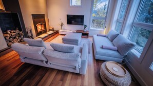 A modern living room with a fireplace, a flat-screen TV, and a large window offering a view of trees. - NEW Modern Mountain Haven | Luxe View, Sauna, Hot Tub & Fireplace (Nelson)