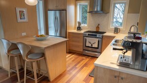 A modern kitchen with wooden cabinets, a stainless steel refrigerator, an oven, and a coffee machine. There are two bar stools and a window providing natural light. - NEW Modern Mountain Haven | Luxe View, Sauna, Hot Tub & Fireplace (Nelson)