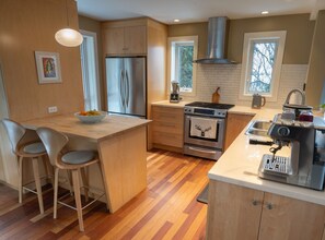 A modern kitchen with wooden cabinets, a stainless steel refrigerator, an oven, and a coffee machine. There are two bar stools and a window providing natural light.