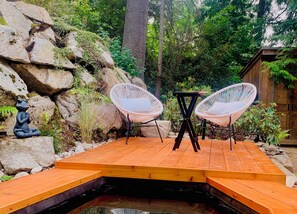 A wooden deck with two white wicker chairs, a frog statue, and a small pond.