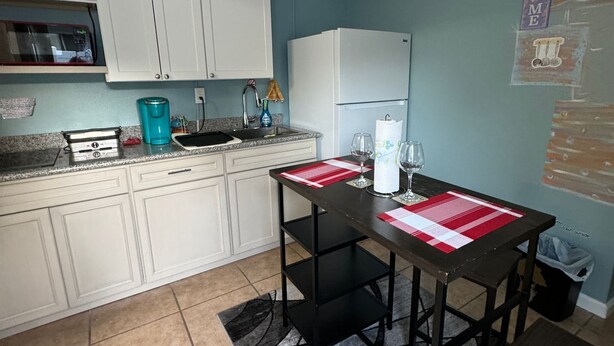 A kitchen with white cabinets, a microwave, a coffee maker, and a refrigerator. There is a dining table with a red placemat and a trash bin. - Neverland. (Jacksonville)