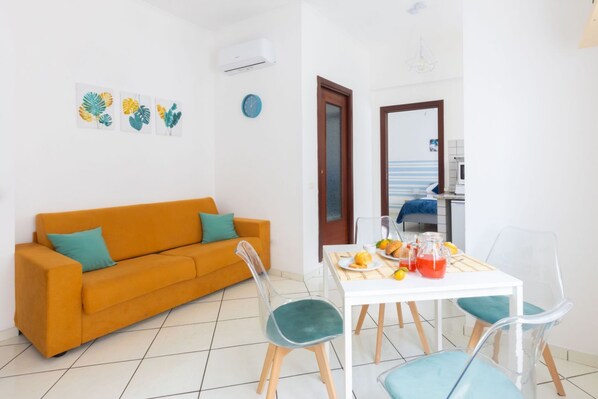 A bright living room with a sofa, chairs, a dining table with breakfast items, and a kitchen area visible through an open door.