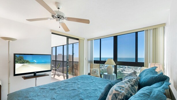 A bedroom with a large bed, a ceiling fan, a TV, and a balcony with a view of the ocean. - Huge Balcony, Amazing Beach Views & Amenities (Bonita Springs)