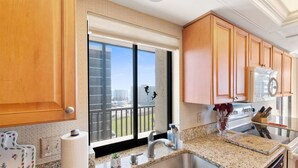 A kitchen with granite countertops, stainless steel sink, wooden cabinets, and a large window with a view of buildings and a balcony. - Huge Balcony, Amazing Beach Views & Amenities (Bonita Springs)