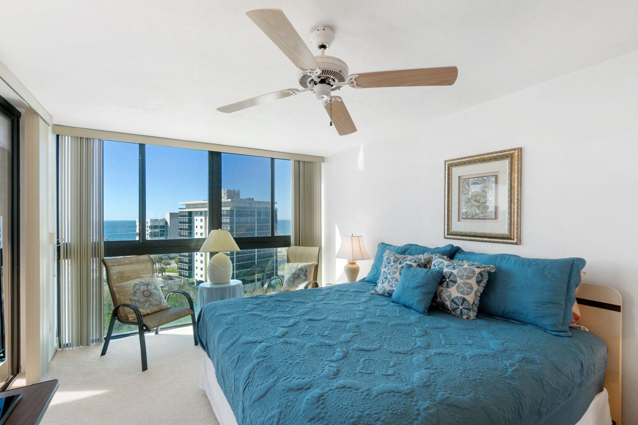 Quarto com uma cama grande, um ventilador de teto, uma janela com vista para edifícios e uma cadeira perto da janela.