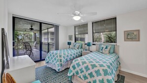 Une chambre avec deux lits, un ventilateur de plafond et un balcon aménagé avec du mobilier d’extérieur.
