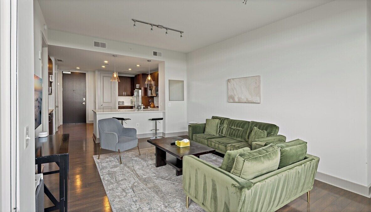 Un salon moderne avec un canapé vert, une table basse et un fauteuil gris. On aperçoit une cuisine au fond.