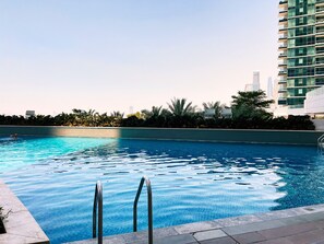 Una piscina con agua azul cristalina, rodeada de una terraza con baldosas y barandillas metálicas. Al fondo, un edificio moderno con múltiples balcones y una hilera densa de palmeras.
