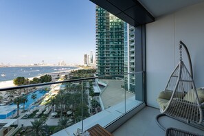Un balcón con vista a una playa y una piscina, una silla colgante y un edificio moderno al fondo.