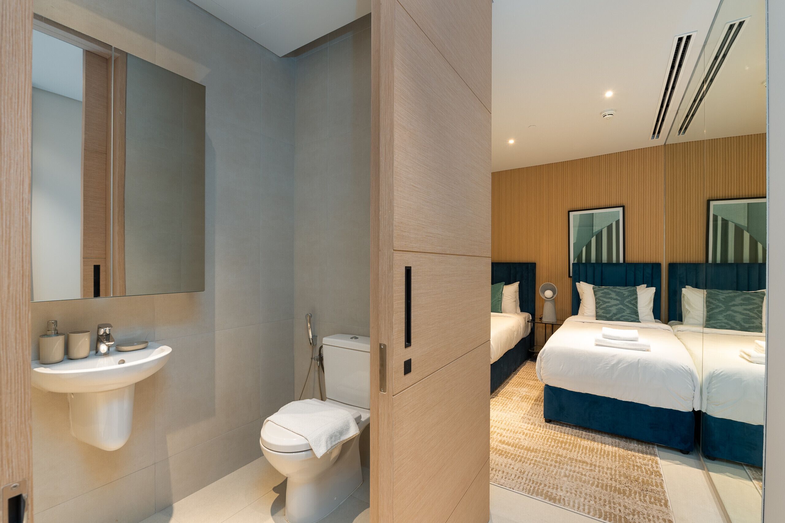Een moderne badkamer met een wastafel, toilet en douche. De kamer heeft een grote spiegel, houten lambrisering en een bed met blauw beddengoed.