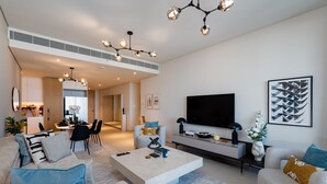 A modern living room with a large flat-screen TV, a coffee table, and a sofa. There is a dining area in the background with a table and chairs.