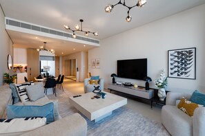 A modern living room with a large flat-screen TV, a coffee table, and a sofa. There is a dining area in the background with a table and chairs.