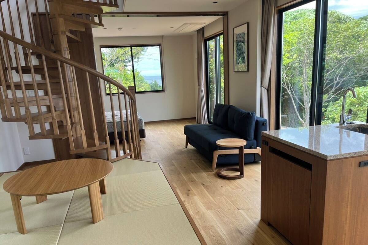 Un salon moderne avec un escalier en bois, un canapé, une petite table ronde et un coin cuisine doté d’un plan de travail.