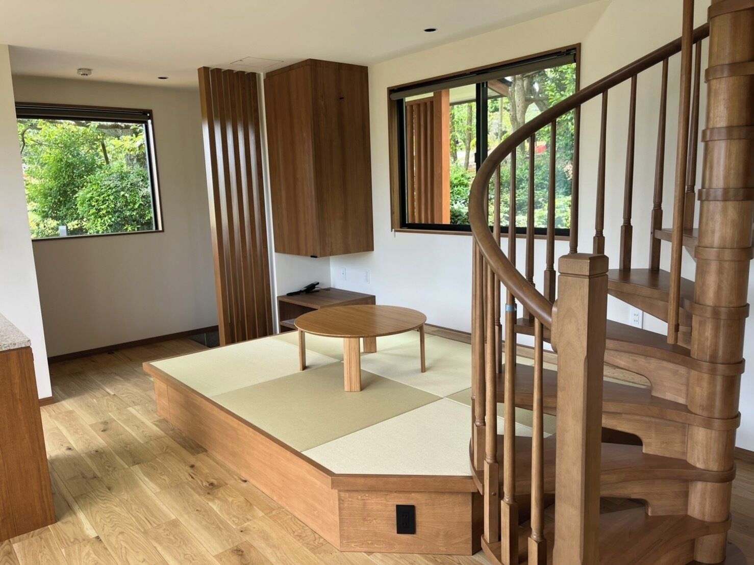 A modern interior with a wooden staircase, a tatami mat seating area, and large windows offering a view of greenery.
