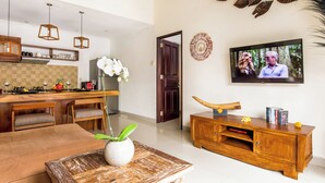 A modern living room with a wooden dining table, a TV mounted on the wall, and a kitchen area in the background. - Villa Joyosa (Seminyak)