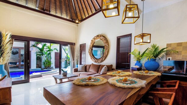 A spacious living area with a wooden dining table, a sofa, and a large mirror. - Villa Joyosa (Seminyak)