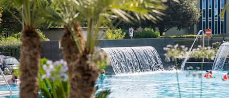 Una piscina con cascada, rodeada de palmeras y un edificio al fondo.